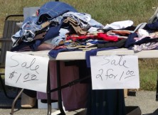 Clothing on a table at a garage sale with sign ‘2 for $1.00’.